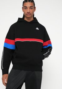 Felpa nera con strisce orizzontali rosse e blu, dettagli bianchi e logo. Materiale morbido, vestibilità comoda, con tasca a marsupio.