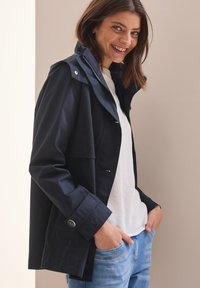 Marinblå vattentät jacka med huva, framdragkedja och knappdetaljer, i kombination med en vit strukturerad topp och ljusblå jeans.