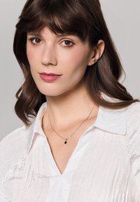 Złoty naszyjnik z czarną zawieszką w kształcie gemu, noszony na teksturowanej białej bluzce z dekoltem w serek. Jasne tło.