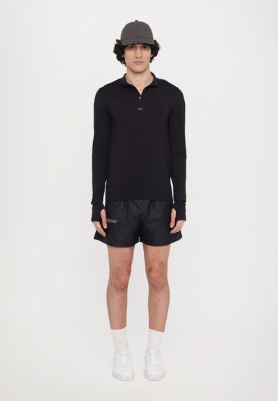 Zwart T-shirt met lange mouwen en een rits tot een kwart, gecombineerd met zwarte sportbroekjes en witte sneakers. Grijze pet en mouwloze handschoenen.