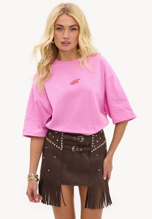 Femme blonde portant un t-shirt rose ample avec un logo rouge et une mini-jupe marron à franges avec des clous argentés et des ceintures à double boucle.