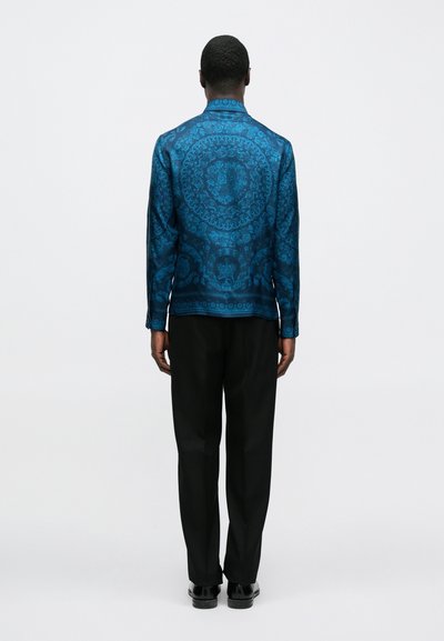 Kék mintás dzseki texturált, fényes felülettel, részletes mintákkal a hátoldalán, fekete nadrággal és cipővel párosítva.