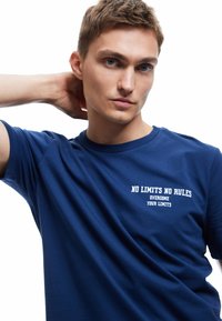 Mørkeblå bomulds t-shirt med rund halsudskæring, korte ærmer og hvid tekst, der siger "INGEN BEGRÆNSNINGER INGEN REGLER OVERKOM DINE BEGRÆNSNINGER."