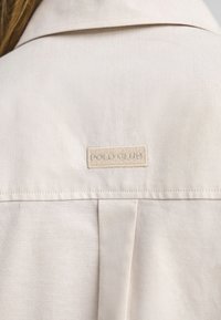 Beżowa koszula z wyraźnym kołnierzykiem i widocznym przeszyciem. Posiada naszytą z tyłu metkę "POLO GLUE", która podkreśla teksturowany materiał.