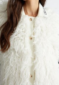 Weißer Kunstfellmantel mit einem strukturierten, lockigen Design, einem cremefarbenen Kragen und Holzknöpfen. Schlichte, lockere Passform mit einem Knopfverschluss.