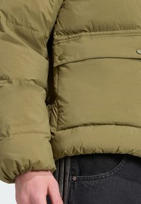 Olīvu zaļš pūķa jakas ar teksturētu apdari, ar kabatu ar vāku, aizdari ar snap pogām un ribotām piedurknēm. Zem jakas redzami melni džinsi.