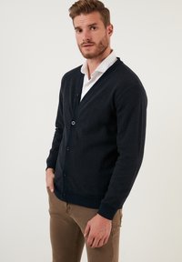 Tumši zila kardigāna jaka, kas izgatavota no mīksta materiāla, ar V veida kaklu, priekšējiem pogām un ribotiem aprocēm un apakšmalu. Reizē ar baltu apkakles krekli.