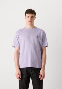 Lavendel bomulds t-shirt med rund halsudskæring og korte ærmer. Har et lille sort trekantet logo på brystet. Par mørke jeans.