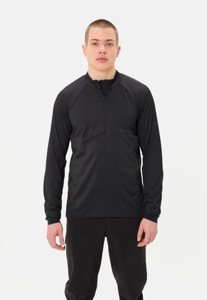 Giovane uomo che indossa un pullover nero a manica lunga con mezza cerniera e pantaloni neri, in piedi davanti a uno sfondo chiaro e uniforme.