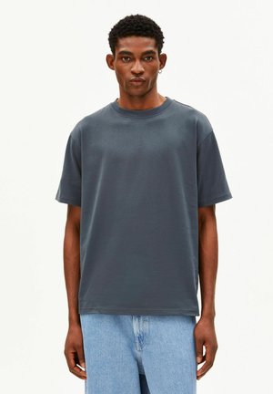 Man met kort kroes haar die een effen donkergrijs oversized T-shirt en lichtblauwe spijkerbroek draagt, staand tegen een witte achtergrond.