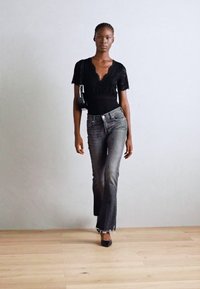 Body en dentelle noire, jeans gris évasés avec des ourlets effilochés, talons noirs. Le modèle pose contre un fond gris clair sur un sol en bois.