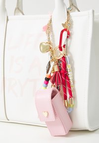 Borsa bianca con finitura testurizzata, decorata con charm colorati e un portachiavi rosa con chiusura a forma di cuore. Dettagli notevoli includono perline e frange.