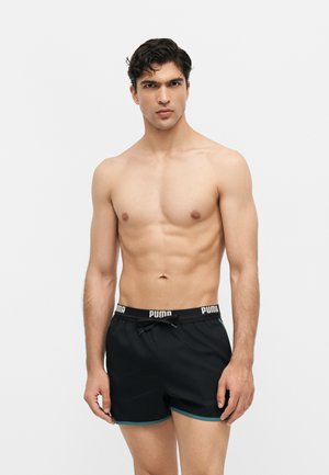 Skjorteløs mand iført sorte Puma badeshorts med turkis kant, stående mod en ensfarvet lysegrå baggrund.