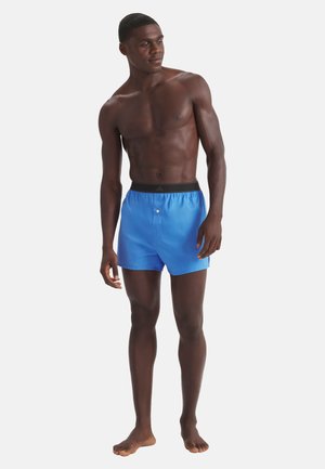 Hombre musculoso de pie, descalzo, vistiendo pantalones cortos azul brillante con cintura elástica negra, mirando ligeramente hacia su izquierda.