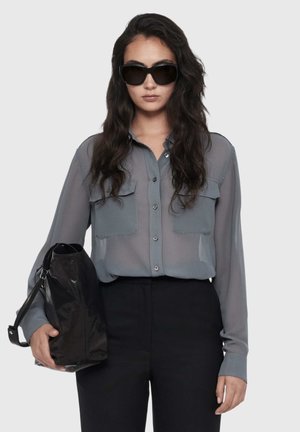 Woman with long dark hair wearing black sunglasses, a sheer gray button-up shirt, black pants, holding a large black bag.