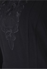 Tessuto nero con ricami floreali scuri nella zona del petto, con una trama liscia e una vestibilità su misura. Dettagli di design sottili arricchiscono l'aspetto complessivo.