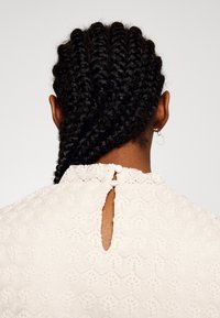 Personne aux cheveux tressés vue de dos, portant un haut blanc texturé avec deux boutons au col et une petite boucle d'oreille.
