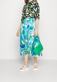 Blusa nera con motivi astratti verdi e bianchi, gonna fluida blu e verde, sandali bianchi e borsa verde con effetto coccodrillo.