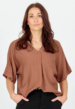 Femme aux longs cheveux bruns portant un chemisier marron ample et un pantalon noir, debout les mains dans les poches devant un fond uni.