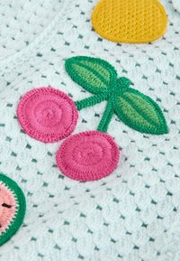 Tela blanca de ganchillo con cerezas rosas bordadas y hojas verdes, parte de un textil colorido con patrón de frutas.
