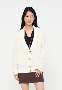 Woman wearing a white textured cardigan with gold buttons over a black top and a brown skirt, standing against a plain white background.