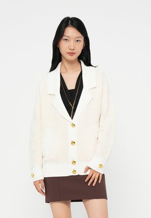 Woman wearing a white textured cardigan with gold buttons over a black top and a brown skirt, standing against a plain white background.