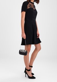 Kvinna i en svart klänning med genomskinlig spets ovanpå, håller i en liten svart clutch med blommotiv, samt svarta högklackade skor.