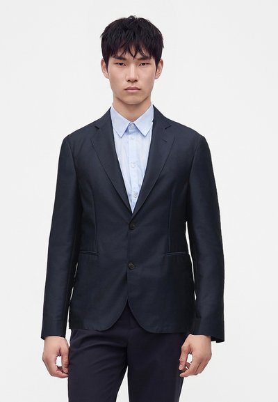Homme portant une veste de costume noire sur une chemise bleue claire boutonnée, debout devant un fond blanc uni.