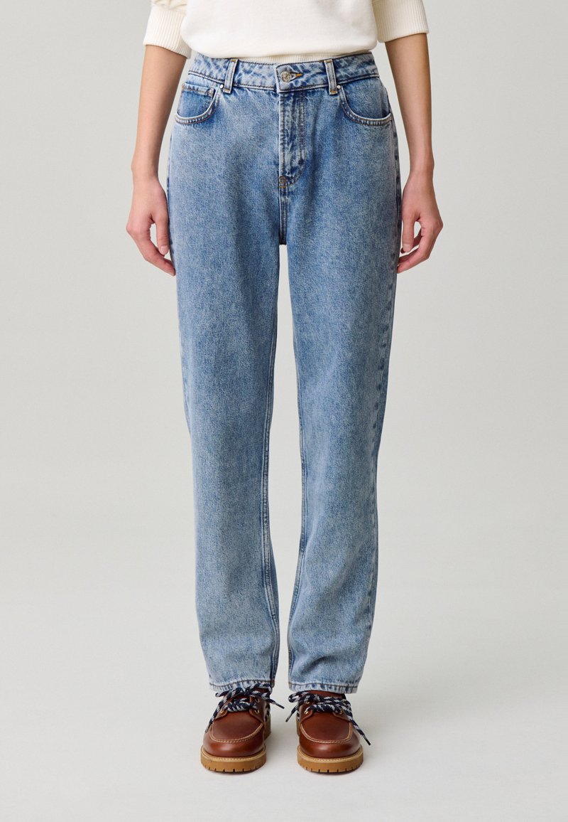 Pantalones vaqueros de mezclilla azul claro con cintura alta, corte relajado, cinco bolsillos y sutiles detalles de desgaste, combinados con zapatos marrones con cordones.