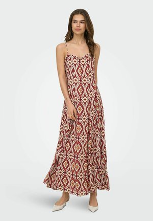 Femme portant une robe sans manches, longue jusqu'aux chevilles, rouge et beige à motifs, avec des fines bretelles et des talons blancs pointus, debout sur un fond uni.