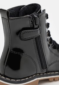 Botín de cuero negro charol con cordones decorativos, cierre con cremallera, forro interior suave y suela de goma con costuras blancas.