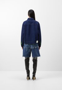 Tummansininen farkkutakki, jossa vihreät korostukset hihoissa, yhdistetty kulutettuihin sinisiin farkkushortseihin ja polven korkeisiin mustiin piikkikoroihin.