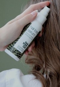 Hårborttagningsserum i en vit flaska med grön lövdesign. Innehåller tea tree-olja. Användare applicerar det på vågigt brunt hår.