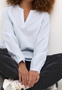 Hellbluse mit Streifen, V-Ausschnitt, langen Ärmeln und Knopfmanschetten. Kombiniert mit dunklen Jeans und weißen Sneakers. Einfaches Design und weicher Stoff.