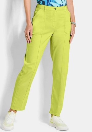 Vrouw draagt fel limegroene casual broek met voorzijde zakken en witte sportschoenen, staand tegen een effen lichte achtergrond.