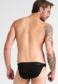 Bikini briefs neri da uomo con finitura liscia, vita elastica e copertura minima, mostrati dal retro con dettagli di tatuaggi visibili.