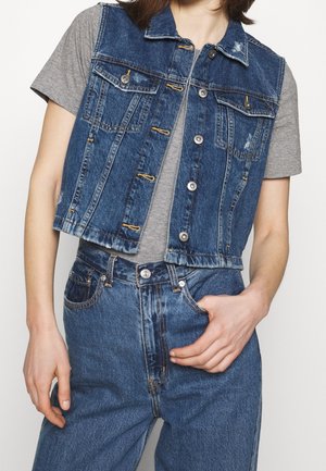 Personne portant un gilet sans manches en jean bleu sur une chemise grise à manches courtes avec un jean bleu en denim taille haute, main dans la poche.