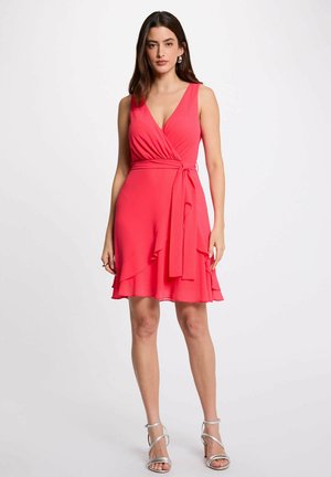 Donna che indossa un vestito avvolgente rosso senza maniche con scollo a V e cintura annodata in vita, abbinato a sandali argentati con tacco alto, in piedi su sfondo bianco.