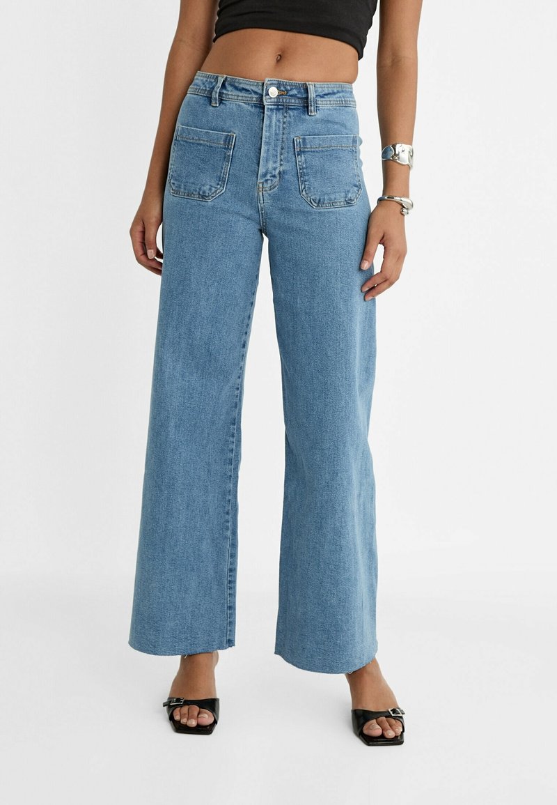 Femme portant un jean taille haute bleu clair à jambes larges avec de grandes poches avant, un crop top noir, des bracelets argentés et des talons ouverts noirs.