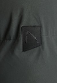 Parche negro en tela verde oscuro con una superficie texturizada. El parche presenta un diseño minimalista de líneas superpuestas.