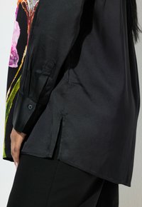 Sort satinbluse med et farverigt blomstermotiv på forsiden, med lange ærmer og sideslids ved kanten.