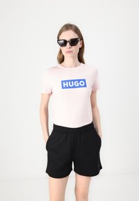Pink cotton t-shirt with blue "HUGO" logo, paired with black elastic waist shorts. Model wears black cat-eye sunglasses.