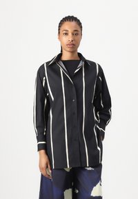Black and white striped button-up shirt with a collar, featuring long sleeves and a relaxed fit. Made of textured cotton fabric.
