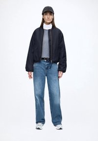 Crna bomber jakna, sivi rolka, svijetloplave široke hlače i tamna kapa. Model stoji na neutralnoj pozadini, noseći bijele tenisice.