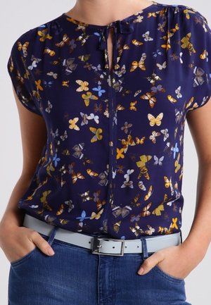 Femme portant un chemisier bleu marine à imprimés papillons multicolores, manches courtes, col noué, jean bleu et ceinture gris clair avec boucle argentée.