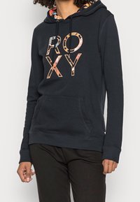 Persona con una sudadera negra con "ROXY" en letras grandes y con un patrón, y un forro interior de capucha floral. Manos relajadas a los lados.