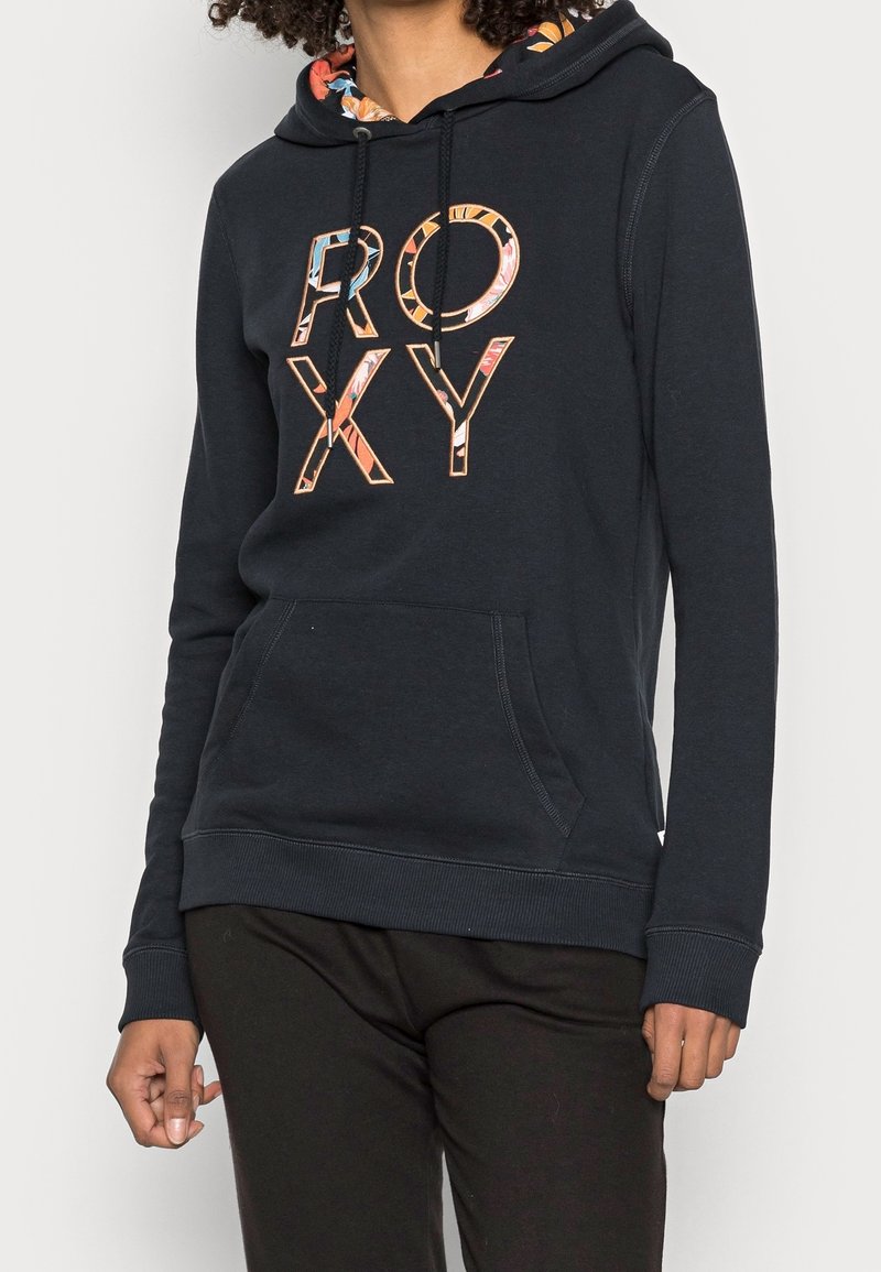 Persona con una sudadera negra con "ROXY" en letras grandes y con un patrón, y un forro interior de capucha floral. Manos relajadas a los lados.