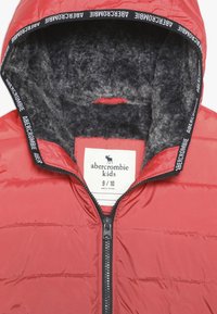 Czerwona kurtka puchowa z pluszowym szarym wykończeniem, z zamkiem błyskawicznym i czarnym logo na kapture. Wewnątrz widoczna metka rozmiaru.