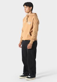 Sudadera color tostado con un bolsillo frontal y un logo, con capucha ajustable. Combinada con pantalones negros de corte holgado y zapatillas negras.