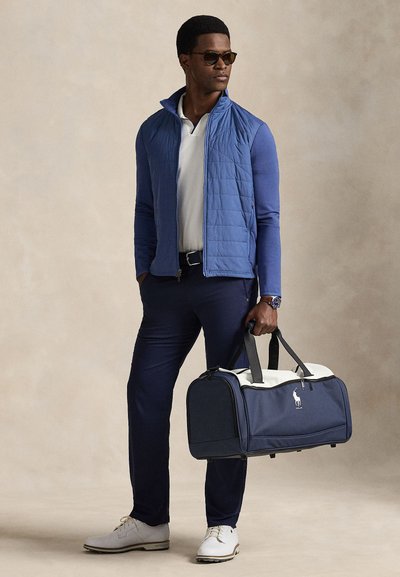 Man som bär solglasögon, blå quiltad väst, vit pikétröja, marinblå byxor, håller en marinblå och vit duffelväska, stående mot en beige bakgrund.